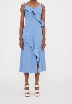 Vestito midi blu senza maniche con dettagli asimmetrici a volant, spalline annodate, indossato con sandali bianchi platform con punta aperta.