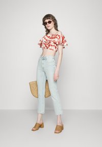 Top cropped tie-dye em vermelho e branco com mangas em babados, jeans de cintura alta em azul claro, sandálias de salto trançadas em tom de castanho e uma bolsa de palha.