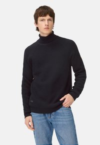 Sort strikket rullekrave-trøje med tekstureret mønster, raglanærmer og ribbet kant. Kombineret med lyseblå jeans.