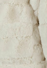 Vit fuskpäls med en fluffig textur och sömmade horisontella paneler i olika nyanser av kräm. Mjuk, pälsliknande utseende.