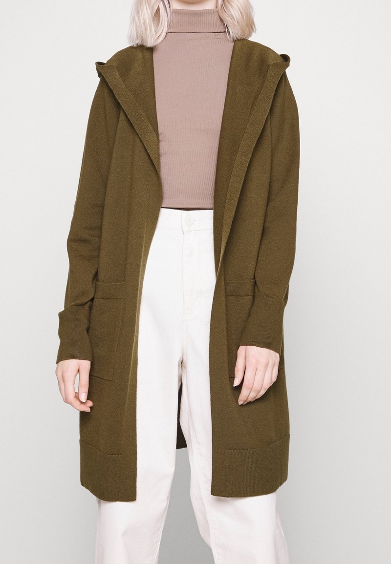 Personne portant un cardigan à capuche vert olive ouvert sur le devant, un col roulé côtelé mauve et un pantalon blanc taille haute, se tenant devant un fond uni.