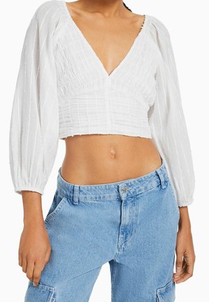 White cropped blouse with a deep V-neck, textured fabric, and puff sleeves. Paired with light blue high-waisted jeans, featuring side pockets.
