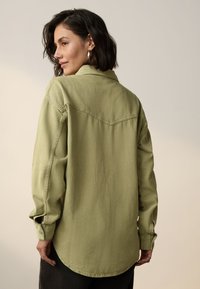 Grünes, übergroßes Hemd mit Knöpfen, langen Ärmeln, Kragen und abgerundetem Saum. Strukturierter Stoff mit markanten Details der Rückennaht.