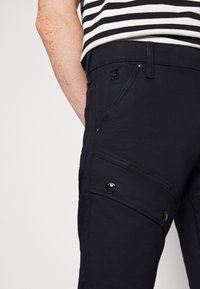 Pantalones casuales negros con un corte ajustado, costuras visibles, bolsillos laterales y botones de metal negro. La tela parece suave y duradera.