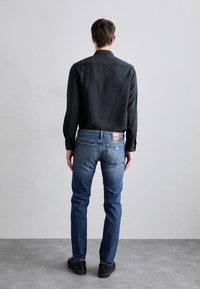 Mörk långärmad skjorta, denimblå jeans med bakfickor och läderlapp. Svarta skor. Sedd bakifrån mot en ljusgrå bakgrund.