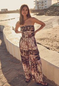 Jumpsuit bez naramenica s vezanjem sprijeda, sa spiralnim uzorkom u nijansama smeđe, kreme i mauve boje. Opušteni kroj s širokim nogavicama.