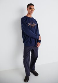 Navy sweatshirt with orange logo, paired with dark pinstriped trousers. Model wearing brown shoes and glasses, arms crossed.