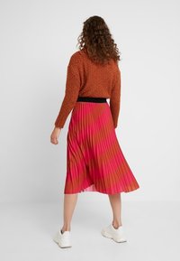 Žena s kudrnatými vlasy má na sobě svetr v barvě rezavé, skládací midi sukni s červenými a růžovými diagonálními pruhy a bílé tenisky, stojí na bílém pozadí.