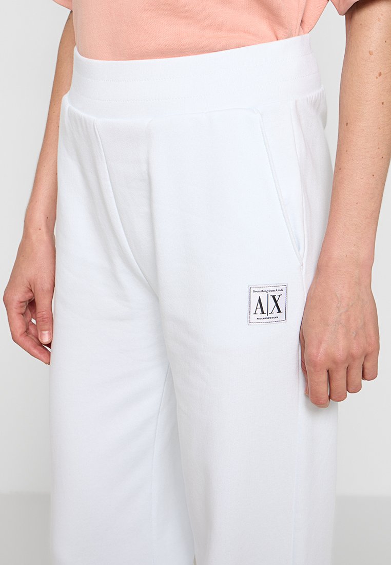 Białe dresy o gładkiej teksturze, z elastycznym pasem i bocznymi kieszeniami. Na wysokości biodra widoczna jest naszywka z logo.