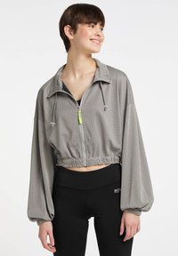 Afklippet, grå atletisk jakke med et mesh-design, snore i kraven og brede ærmer, parret med sorte leggings.