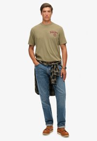 Camiseta de algodón verde oliva con estampado en el pecho, jeans de mezclilla azules, camisa de cuadros atada a la cintura, zapatos marrones con cordones y un reloj.