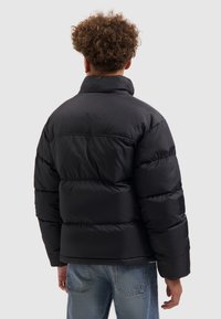 Zwarte pufferjack met een hoge kraag, gequilte secties en een glanzende afwerking. Draag het over lichtblauwe jeans, gezien van achteren.