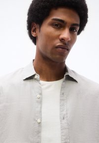 Νεαρός με κοντά σγουρά μαλλιά που φοράει ανοιχτό γκρι πουκάμισο με κουμπιά πάνω από λευκό μπλουζάκι, κοιτάζοντας κατευθείαν μπροστά αντικρίζοντας λευκό φόντο.