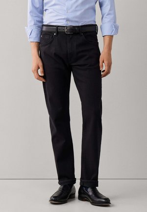 Man draagt een zwarte broek, zwarte riem, zwarte loafers en een lichtblauw gestreept overhemd met opgerolde mouwen.