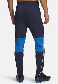 Marineblauwe sportbroek met blauwe meshpanelen, elastische tailleband, slanke pasvorm en reflecterende accenten langs de naden. Er worden zwarte voetbalschoenen gedragen.
