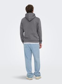 Man met een grijze hoodie, lichtblauwe spijkerbroek en beige schoenen, staand en met de rug naar de camera tegen een effen lichte achtergrond.