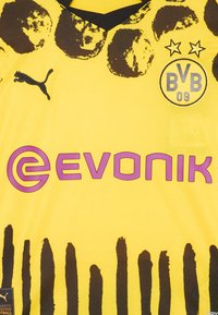 Maglia da calcio gialla con motivi neri, caratterizzata da un logo Puma nero, un distintivo rotondo del club e il logo rosa "Evonik" sulla parte frontale.