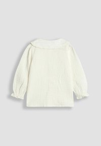 Blusa de bebé de manga larga blanca con tejido arrugado, cuello Peter Pan con volantes y puños fruncidos sobre un fondo gris claro.