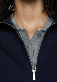 Granatowa kurtka na zamek z szarym, guziczkowym kołnierzem. Tkanina wydaje się gładka, z metalowym zamkiem błyskawicznym i szarymi guzikami.