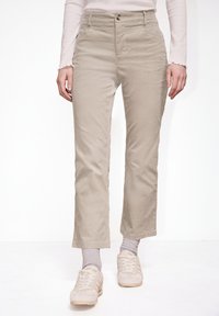 Personne portant un pantalon en velours c ôtelé beige, des chaussettes gris clair et des baskets couleur crème, debout devant un fond blanc.