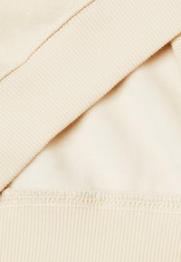 Tessuto di felpa color crema che mostra dettagli a costine sul colletto e sull'orlo inferiore, con una texture interna liscia e cuciture visibili.