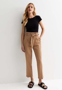 Haut noir à manches courtes, pantalon beige taille haute avec une ceinture à nouer, et sandales noires avec un accent doré. Tissu lisse, coupe ajustée.