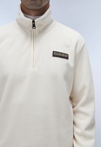 Hvid fleece pullover med halv-lynlås krave. Har et sort logo-patch på venstre bryst. Blød tekstur, afslappet design.