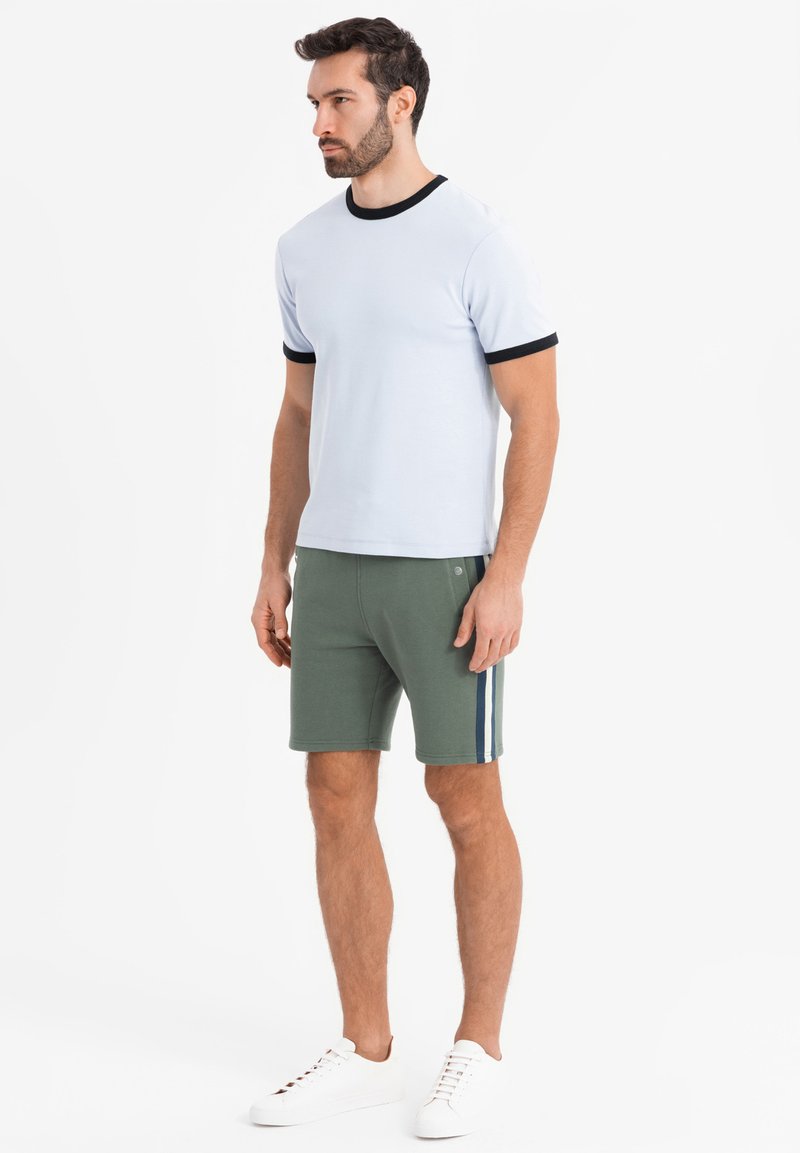 Homme debout portant un t-shirt blanc avec une bordure noire, un short vert avec des bandes latérales et des baskets blanches sur fond blanc uni.