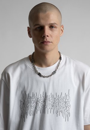 Noor, kellel on ajatud pea, kannab valget graafilist T-särki ja paksu kettkaelakeed ühetaustalise taustaga.