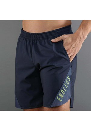 Hombre que lleva pantalones cortos deportivos azul oscuro con cinturilla elástica y la palabra "ENDLESS" impresa en amarillo en el lateral, mano en el bolsillo.