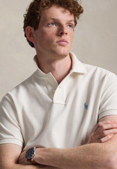 Ανδρας με κόκκινα μαλλιά και φακίδες, φοράει εκρού πόλο μπλούζα και καφέ δερμάτινο ρολόι χειρός, κοιτάζοντας ελαφρώς προς τα δεξιά πάνω σε μονόχρωμο φόντο.