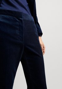 Granatowe spodnie kordowe o prążkowanej fakturze, z płaskim przodem i kieszeniami bocznymi. Zbliżenie pokazuje pas i umiejscowienie rąk.