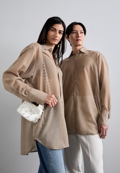 Camicie beige con bottoni, a righe verticali, maniche lunghe e vestibilità rilassata; un modello tiene in mano una piccola borsa bianca con perle.