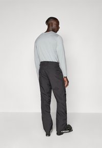 Pantalons de neige noirs avec une finition mate, dotés de passants de ceinture et de poches latérales, associés à un t-shirt à manches longues gris. Bottes de ski visibles.