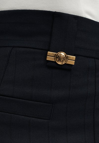 Μαύρο ραμμένο παντελόνι με χρυσό διακοσμητικό κουμπί που έχει κεφάλι Μέδουσας στη μέση, συνδυασμένο με λευκό τοπ.