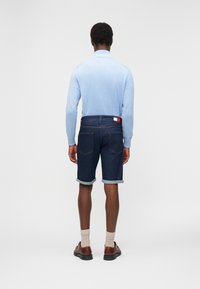 Άνδρας που φορά ανοιχτό γαλάζιο πουκάμισο με μακριά μανίκια, σκουρό μπλε τζιν σορτς γυρισμένα, με ετικέτα λογότυπου, μπεζ κάλτσες και καφέ δερμάτινα παπούτσια, στέκεται με την πλάτη προς το μέρος μας.