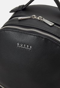 Sac à dos en cuir texturé noir avec quincaillerie argentée, deux compartiments zippés et une plaque logo indiquant "GUESS EST. 1981."