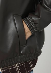 Veste en simili cuir noire avec une taille et des poignets côtelés, dotée d'une poche latérale et d'une texture lisse. L'arrière-plan présente un motif à carreaux.