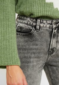 Suéter de punto verde con un patrón texturizado, combinado con jeans grises desteñidos que tienen una cinturilla negra y detalles en el bolsillo frontal.