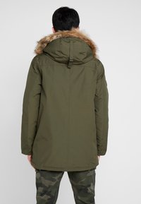 Parka verde oliva con capucha de pelo, con un corte recto y una textura suave, llevada sobre pantalones de camuflaje.