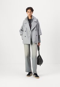 Manteau gris croisé avec de grands revers, coupe oversize et boutons noirs. Associé à un jean bleu clair et des baskets noires. Accessoire : sac noir.