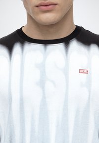 Άνδρας με μαύρο-άσπρο πουκάμισο tie-dye με μικρό κόκκινο λογότυπο Diesel στο στήθος, εμφανιζόμενος από τον λαιμό μέχρι τον άνω κορμό.