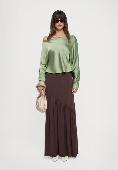 Grøn satin off-shoulder top, brun maxi nederdel med diagonal plissé, holder en beige clutch og bærer solbriller, stylet med spidse beige sko.