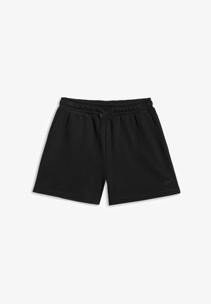 Jeune personne aux longs cheveux foncés portant un sweat à capuche noir, un short noir, des chaussettes blanches et des baskets noires et blanches, debout les mains dans les poches.