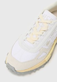 Witte sportieve schoen met details van suède en mesh, crèmekleurige veters, gele accenten op de zool en een gestructureerde grijze rubberen buitenzool.