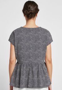 Kvinde med mørkt hår i en knold iført en kortærmet marineblå bluse med et hvidt abstrakt prikket mønster og peplumkant.