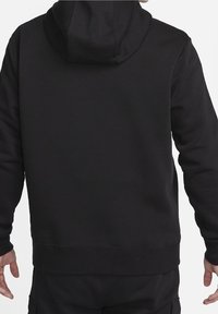 Mann trägt einen schlichten schwarzen Hoodie und passende schwarze Hose, gezeigt von hinten mit entspannt an den Seiten hängenden Händen vor weißem Hintergrund.
