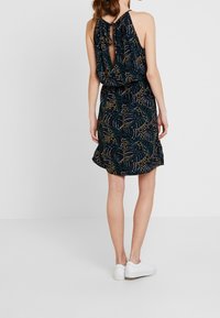 Robe sans manches en tissu sombre, ornée d'un motif de feuilles vertes et dorées. Le détail à l'arrière comprend une ouverture en forme de clé fermée par un cordon.