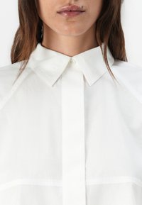 White button-up shirt made of lightweight fabric, featuring a classic collar and a structured design with visible seam details.