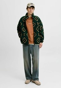 Grüne gemusterte Fleecejacke, braunes Hemd, blaue Jeans, beiger Schuhe und eine hellbraune Mütze. Die Jacke hat geometrische Designs.